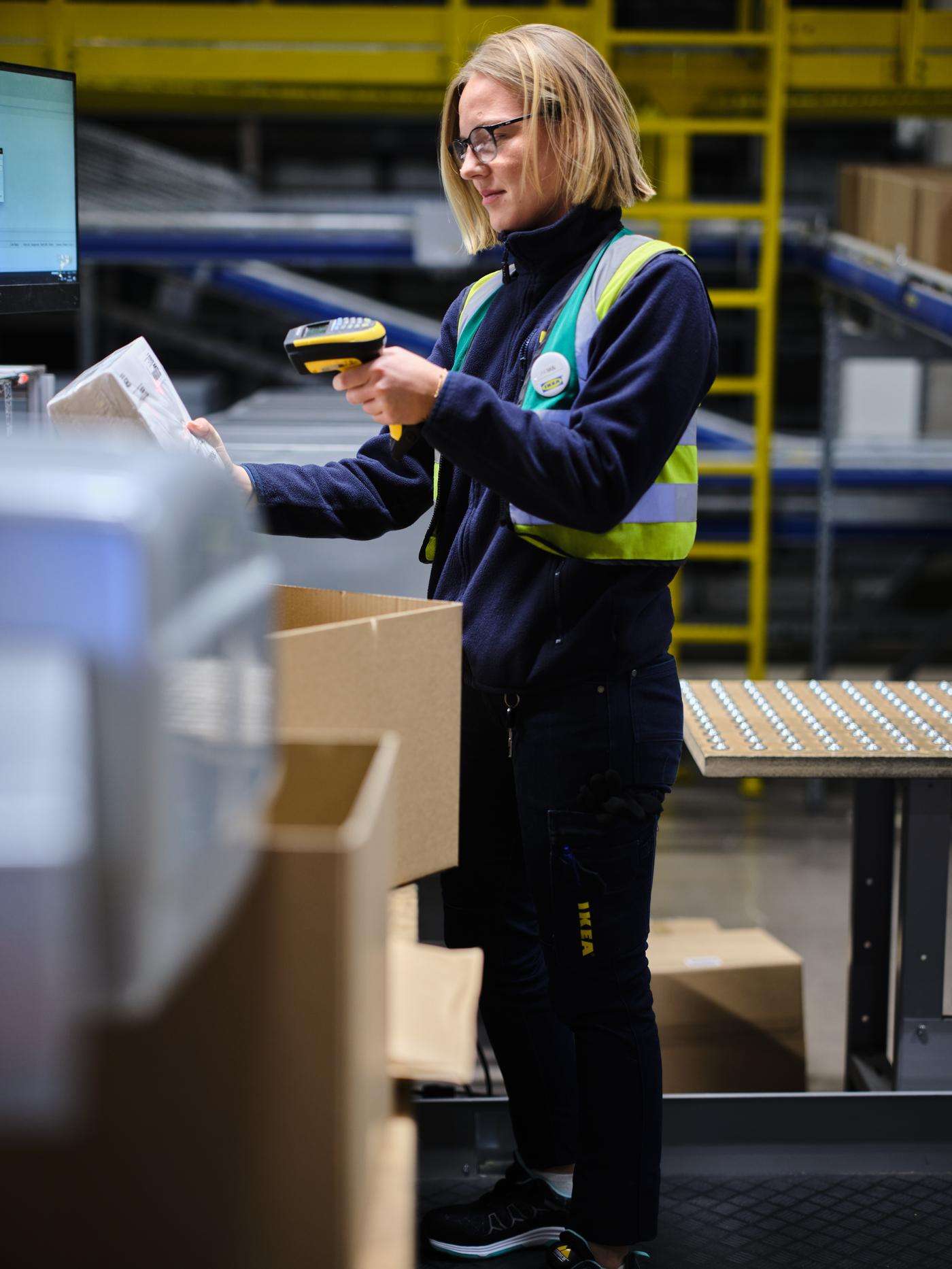 En IKEA medarbetare står i ett lager och skannar ett paket. I förgrunden syns staplade kartonger.