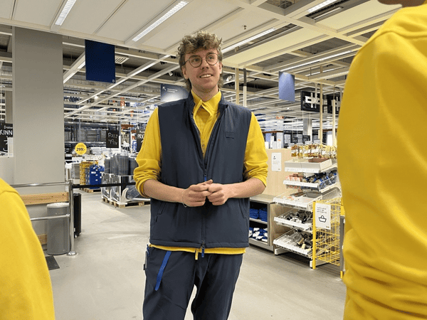 En IKEA medarbetare står i saluhallen i ett varuhus. I förgrunden syns axlarna av två andra medarbetare.