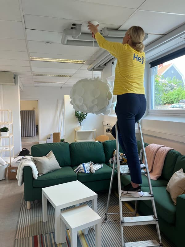 En IKEA medarbetare står på en stege och hänger upp en lampa i taket på Stadsmissionen i Västerås. I bakgrunden syns en stor grön soffa.