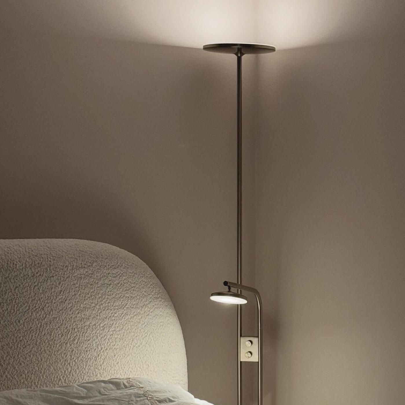 En ISAJKT golvlampa med både uppåtriktat och nedåtriktat ljus står intill en säng med ljus textil i ett mjukt belyst sovrum.