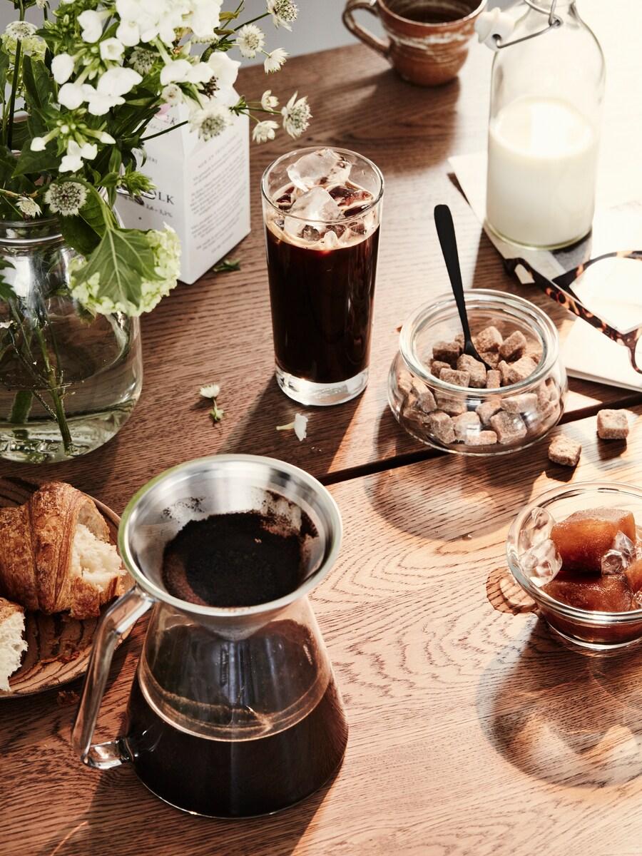En iskaffe förbereds på ett bord och omkring står en vas med blommor, en burk med sockerbitar och en kaffebryggare.