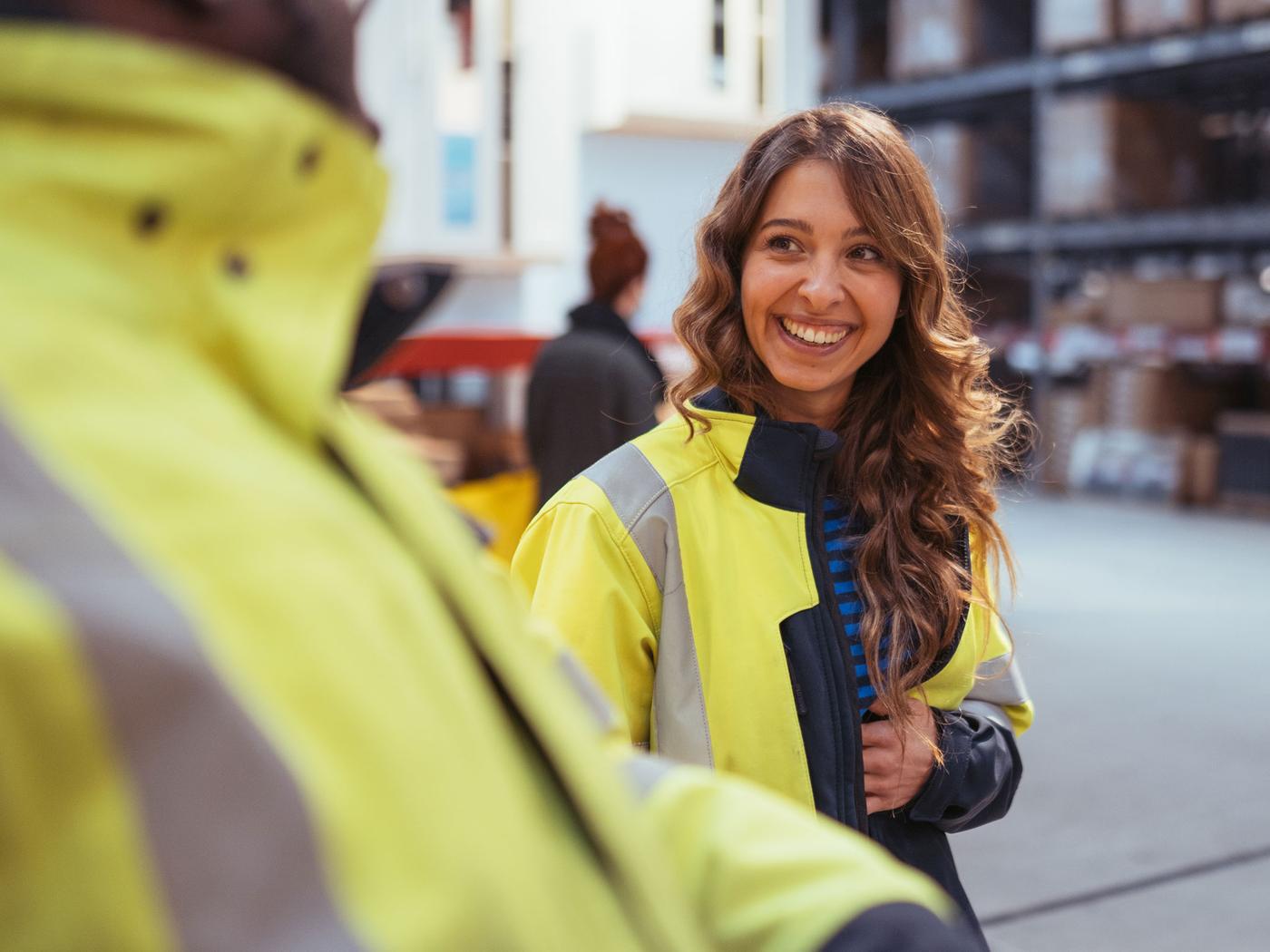 En kvinde i en gul jakke taler med en mand i et lagerlokale, omgivet af kasser og reoler.