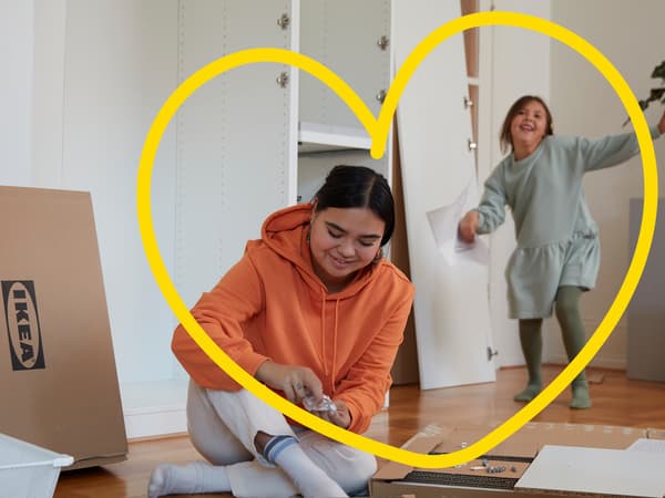 En kvinde i orange trøje sidder på gulvet og samler et IKEA møbel, mens hendes datter danser rundti baggrunden.