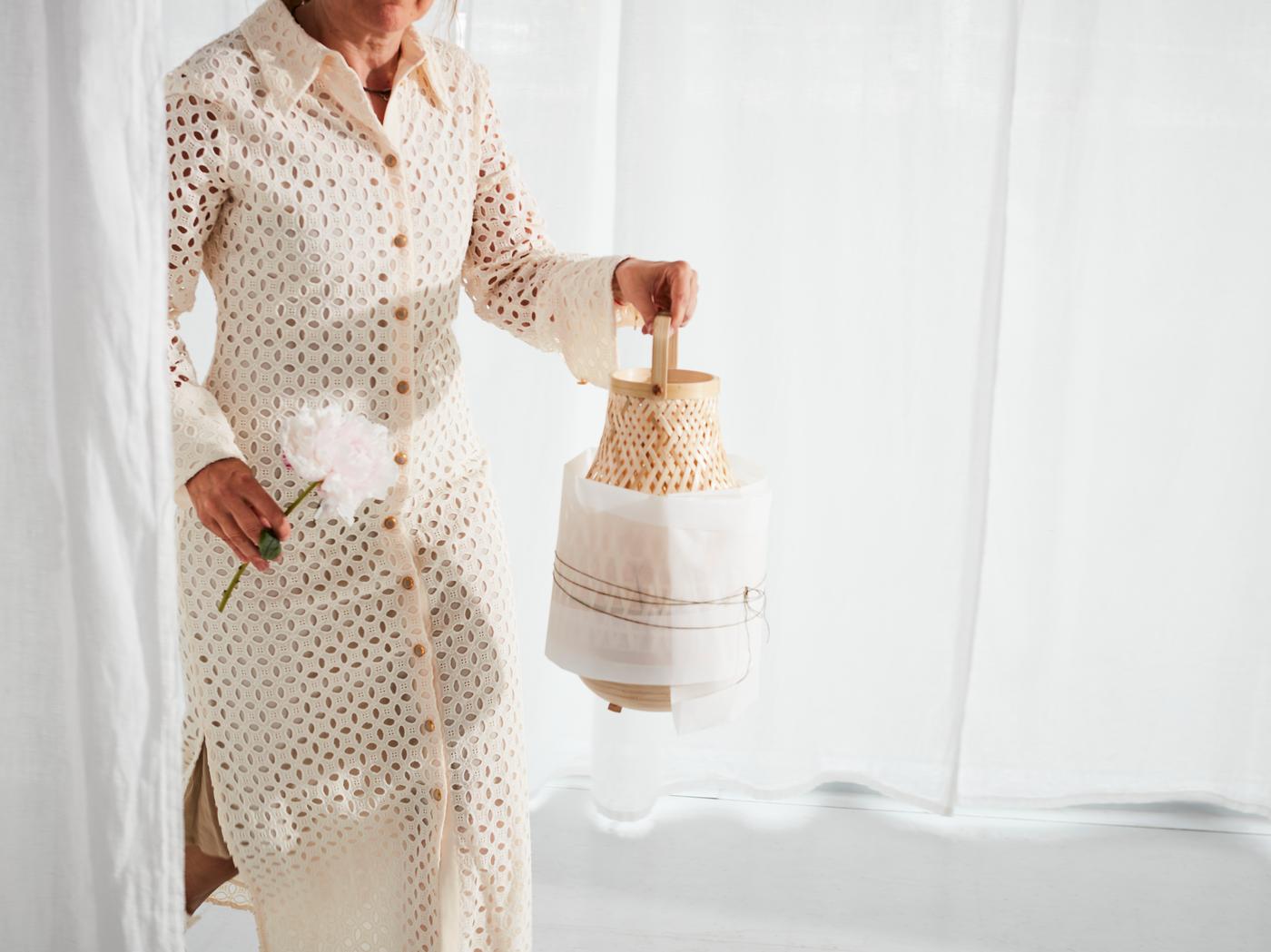 En kvinde klædt i en beige kjole bærer en MISTERHULT bordlampe i vævet bambus, der er delvist indpakket i papir.