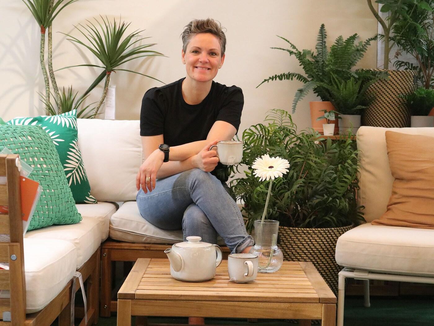 En kvinde sidder i en NÄMMARÖ modulopbygget havesofa med hvide hynder og grønne puder omgivet af grønne planter.