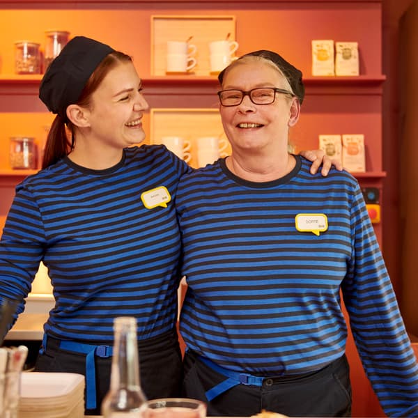 En kvindelig IKEA medarbejder holder armen om en ande kvindelig IKEA medarbejder de smiler begge to og står foran en IKEA udstilling.