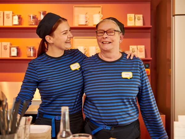 En kvindelig IKEA medarbejder holder armen om en ande kvindelig IKEA medarbejder de smiler begge to og står foran en IKEA udstilling.