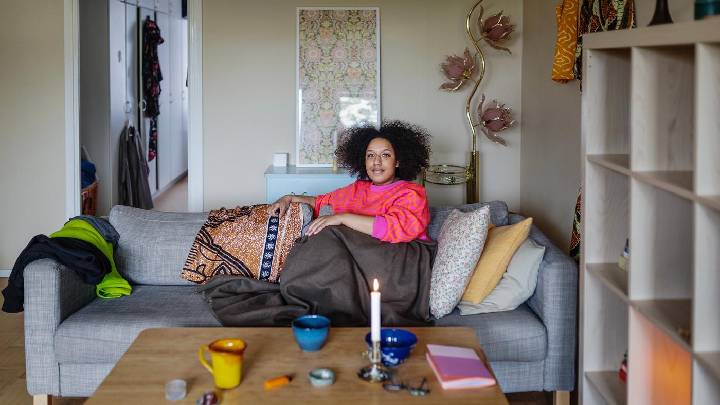 En kvinna i rosa och röd tröja sitter ihopkurad i en grå soffa. Hon har en filt över benen. Soffan står i ett vardagsrum. 
