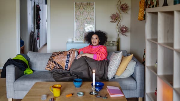 En kvinna i rosa och röd tröja sitter ihopkurad i en grå soffa. Hon har en filt över benen. Soffan står i ett vardagsrum. 
