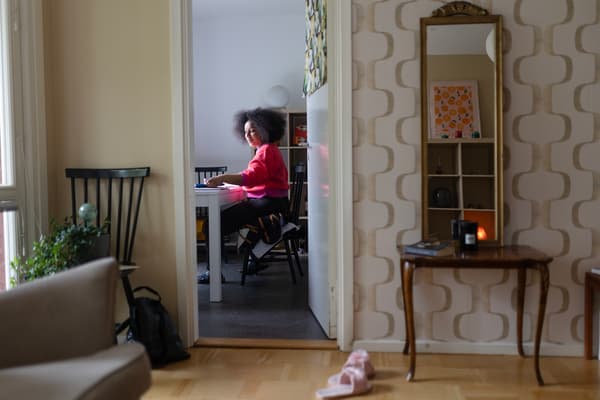 En kvinna i rosa och röd tröja sitter vid ett skrivbord i ett arbetsrum. Utanför rummet står ett par rosa innetofflor. 