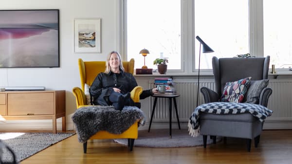 En kvinna i svart klänning sitter i en gul fåtölj i ett vardagsrum. Hon sträcker sig efter en mugg som står vid ett bord bredvid fåtöljen.