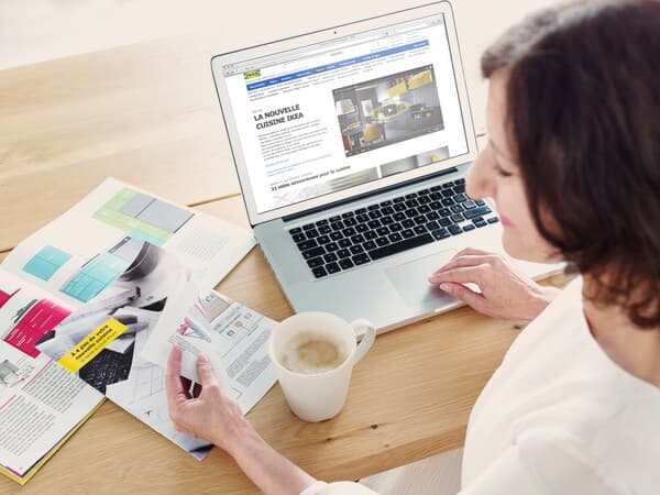 En kvinna sitter vid ett köksbord men en dator och IKEA broschyrer framför sig.
