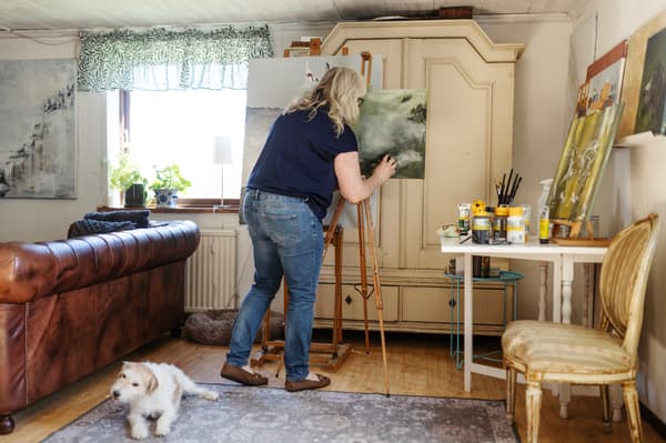 En kvinna står och målar på en tavla som står på ett staffli. På golvet bakom henne ligger en vit hund. 
