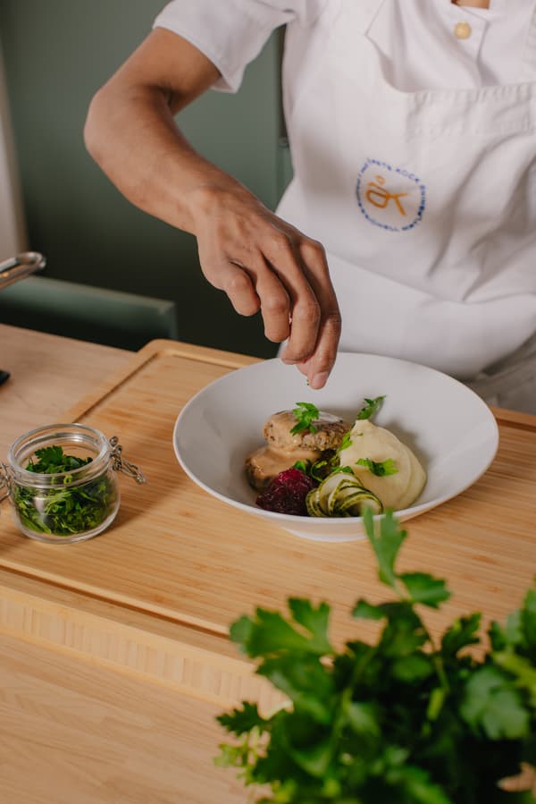 En kvinna strör hackad bladpersilja över en tallrik med potatismos, pannbiff, gräddsås, lingon och pressgurka.