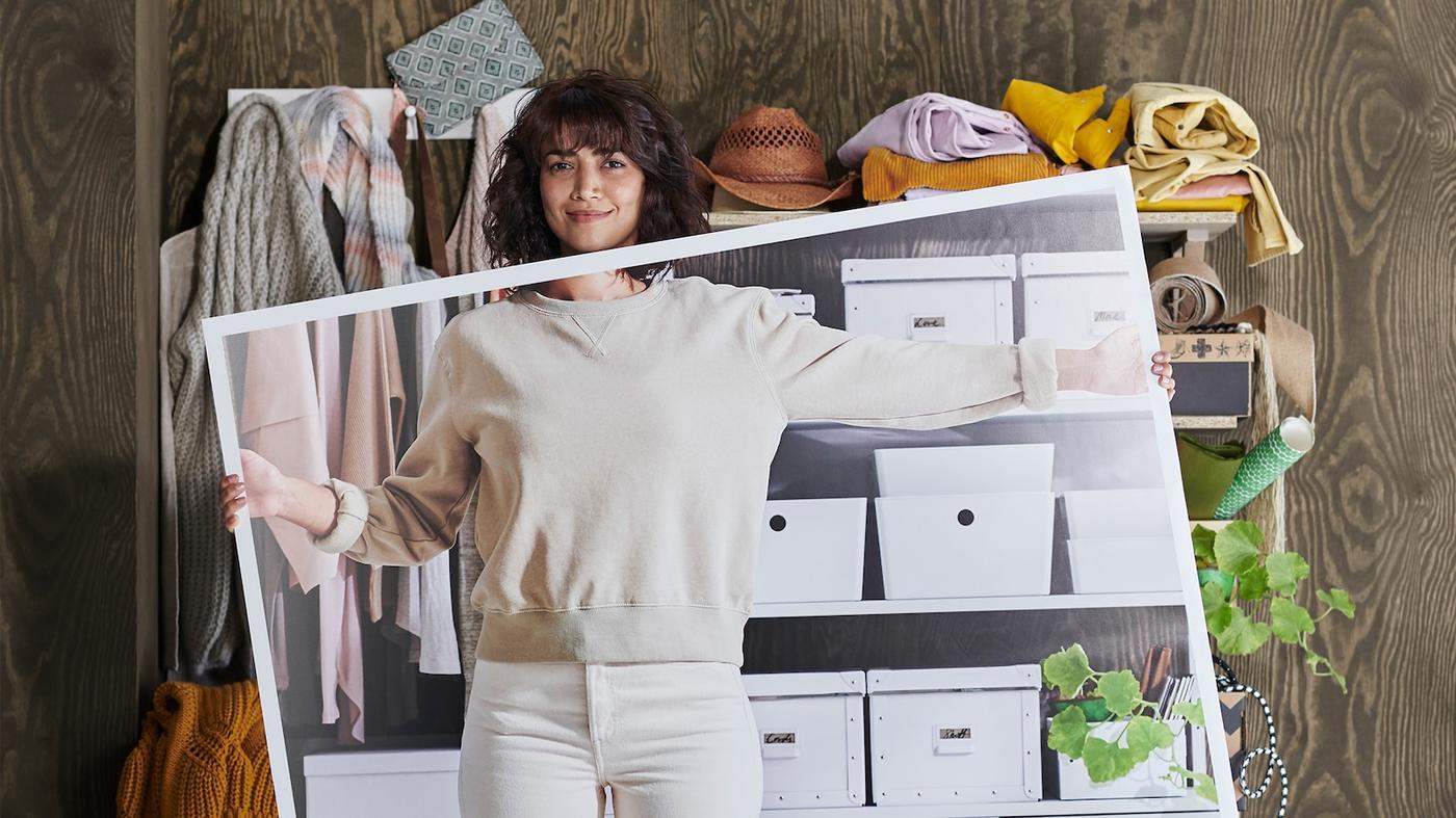 En kvinne i hvite klær står foran hyller fulle av rot. I hendene har hun et stort bilde av hvordan hyllene ser ut når de er ryddige.