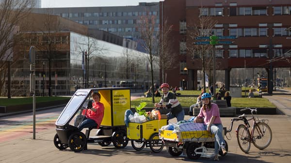 En lastcykel med tak, en lastcykel och ett cykelsläp står parkerade framför en brun byggnad.