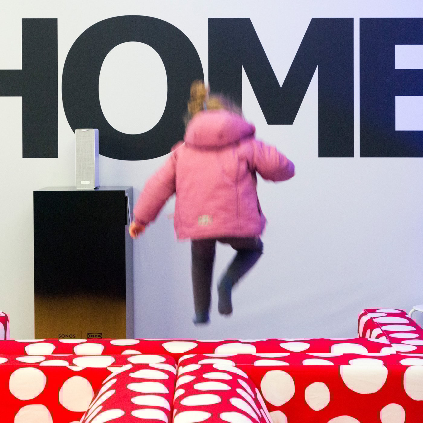 En lille pige i en lyserød jakke hopper i en rød sofa med hvide prikker i baggrunden står home på væggen.