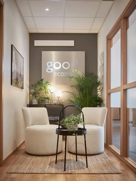 En liten, modern vänthörna med två vita stolar, ett runt svart bord med en krukväxt och texten 'good economy' på väggen bakom. Gröna växter och glaspartier kompletterar inredningen.