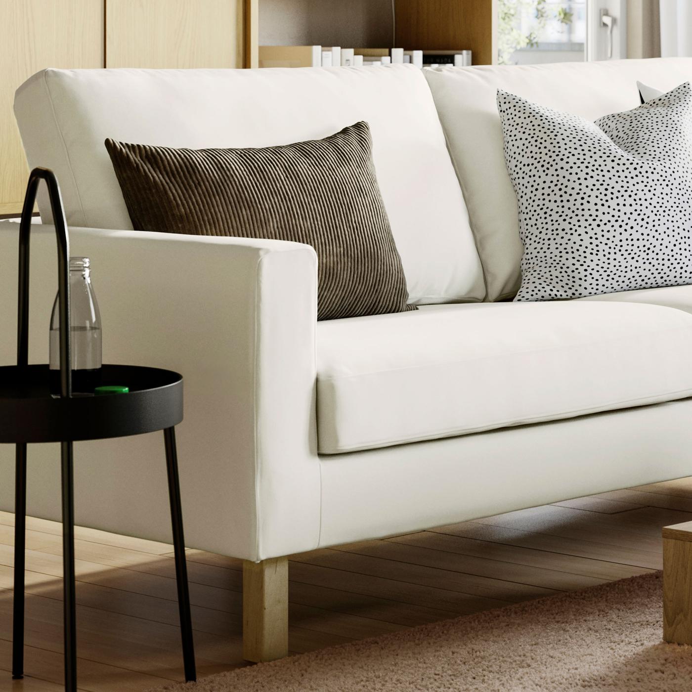 En ljus SALTSJÖBADEN soffa men en brun och en vit- och svartprickig kudde i står intill en vitt litet sidobord framför ett skåp i trä. 