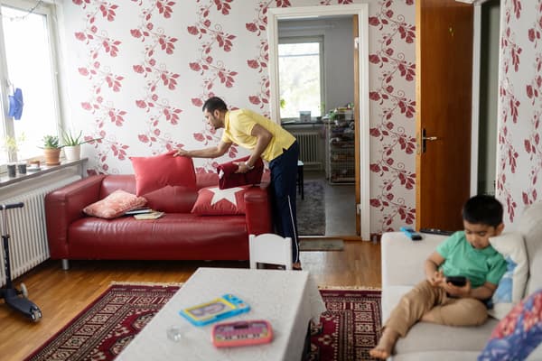 En man fixar soffkuddarna i en röd soffa. I en soffa bredvid sitter ett barn och spelar. 
