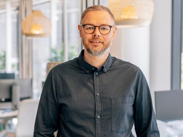 En man i grå skjorta, skägg och glasögon står i en kontorsmiljö.