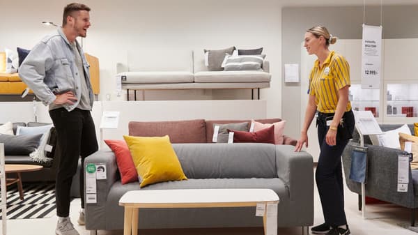 En man klädd i jeansjacka och en kvinna klädd i IKEA unfirom pratar med varandra. På bilden syns en grå soffan med en röd och en gul kudde i.