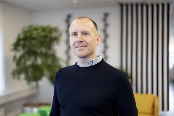 En man med kort hår, skjorta och en mörk tröja står i ett kontorslandskap.