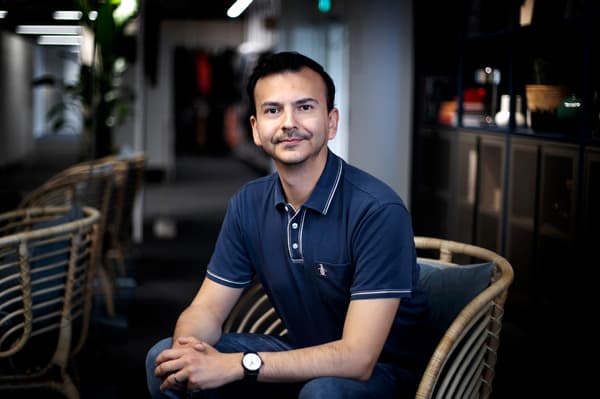 En man med mörkt hår och blå pikétröja sitter framåtlutad i en fåtölj med händerna knäppta och ler mot kameran.