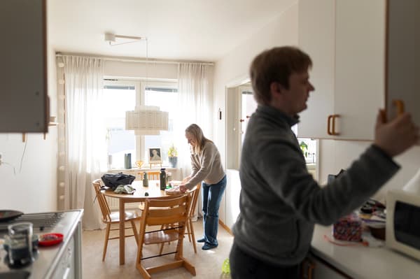 En man och en kvinna står i ett kök. Mannen öppnar en kökslucka och kvinnan står vid ett köksbord och förbereder mat. 