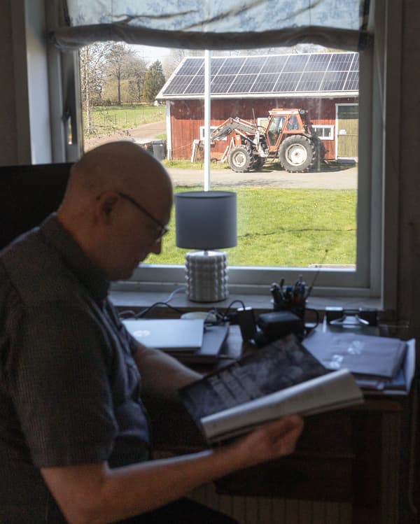 En man sitter vid ett fönster och läser. Utanför fönstret står en röd traktor parkerad. 