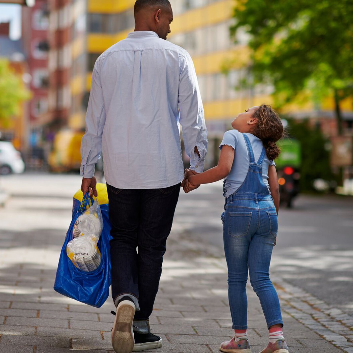En mand går med ryggen til og holder en lille pige i hånde mens han holder en blå IKEA bærepo i den anden hånd.
