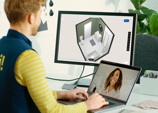 En mand i ikea-uniform sidder ved en computer. På en lille skærm ser han en kunde og på en stor skærm har han et billede af et badeværelse.