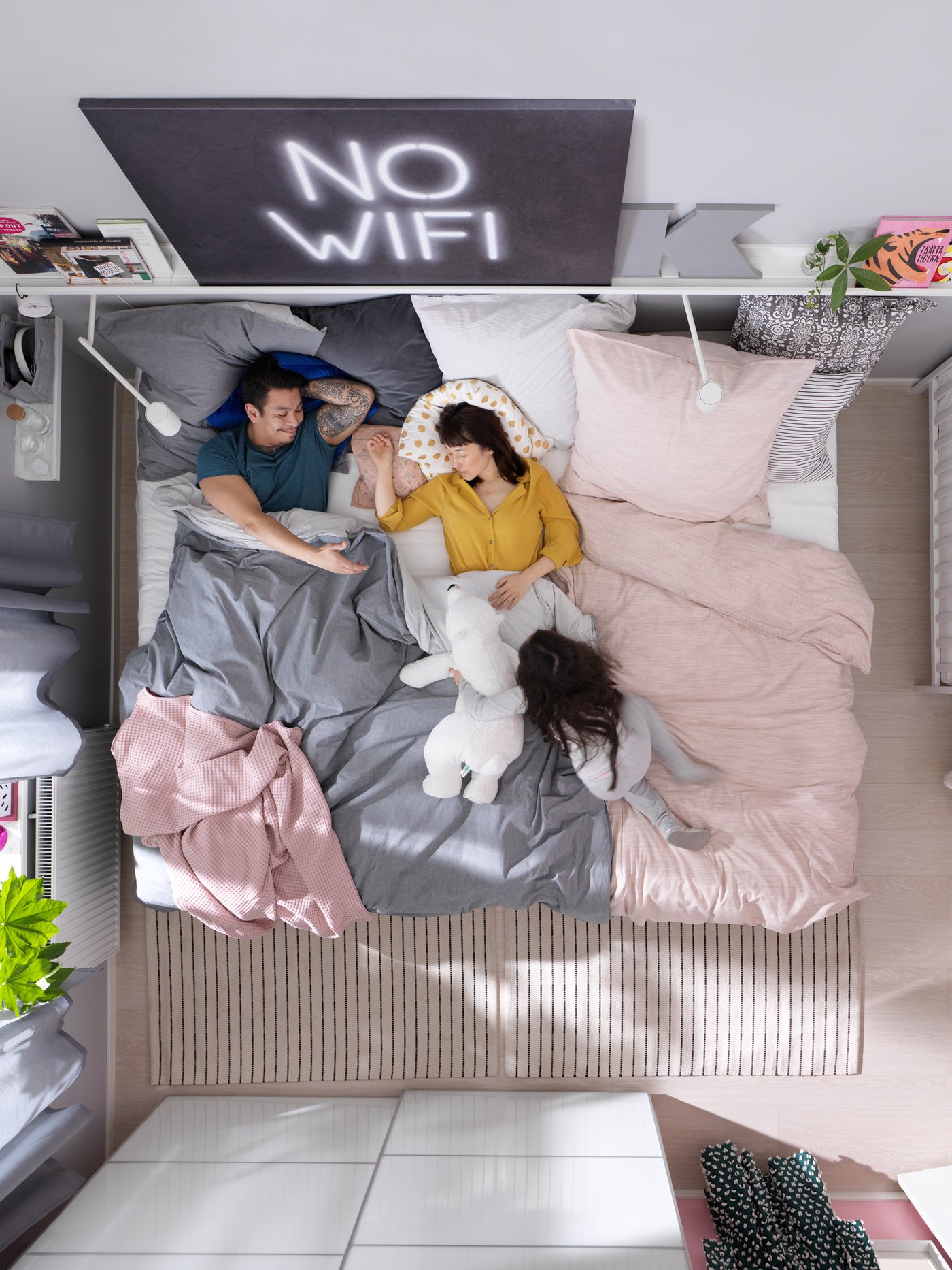 En mand og en kvinde ligger i en seng i et soveværelse i sengen er der to tøjdyr og over sengen hænger et sort skilt med teksten no wifi.