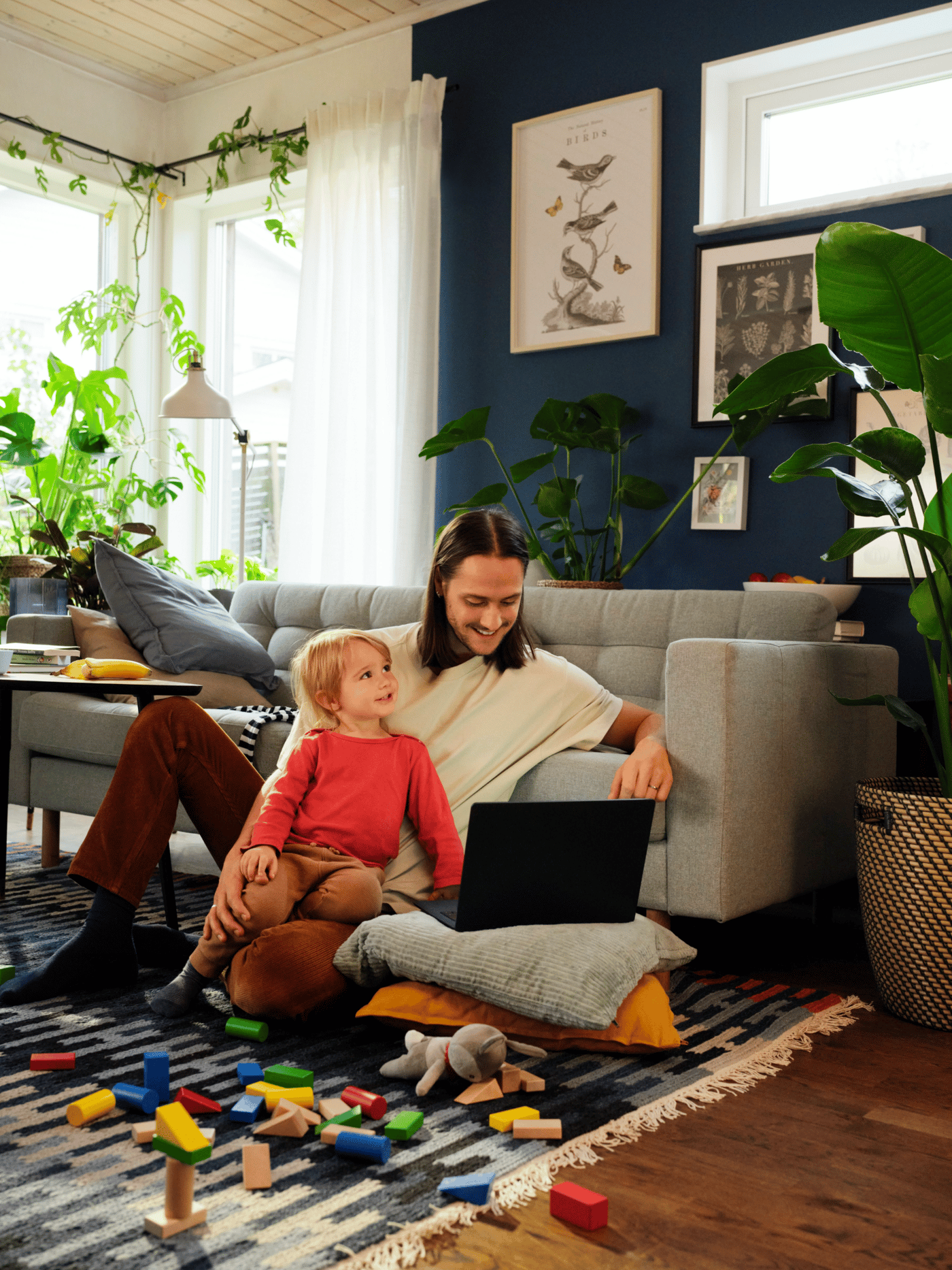 En mand ser på en computer sammen med et barn, de sidder på gulvet i en stue foran dem ligger legetøj spredt ud på gulvtæppet.