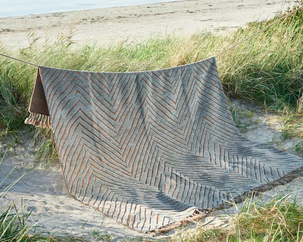 En matta med sicksackmönster i grönt bått hänger mellan två stolpar vid sandstrand och gräs i solnedgång.