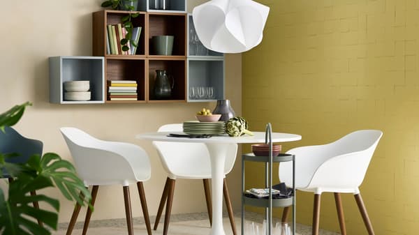 En modern matplats med ett runt vitt bord, fyra vita stolar och en hylla med tallrikar och glas på väggen. En vit lampa hänger ovanför bordet.
