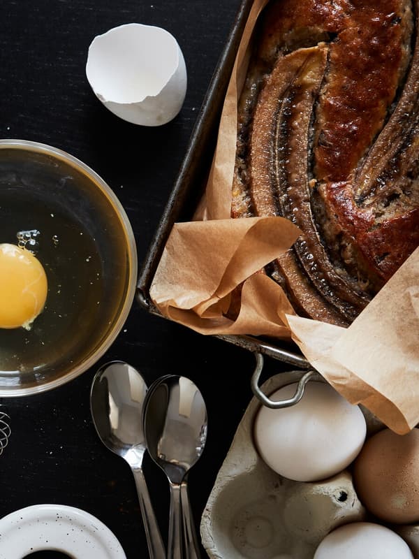 En närbild på ett nybakat bananbröd och ingredienser som ägg och bakredskap ligger kvar framme.