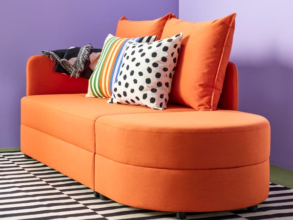 En orange LINDÅKRA bäddsoffa mot en lila bakgrund och en svartvit matta under bäddsoffan. 
