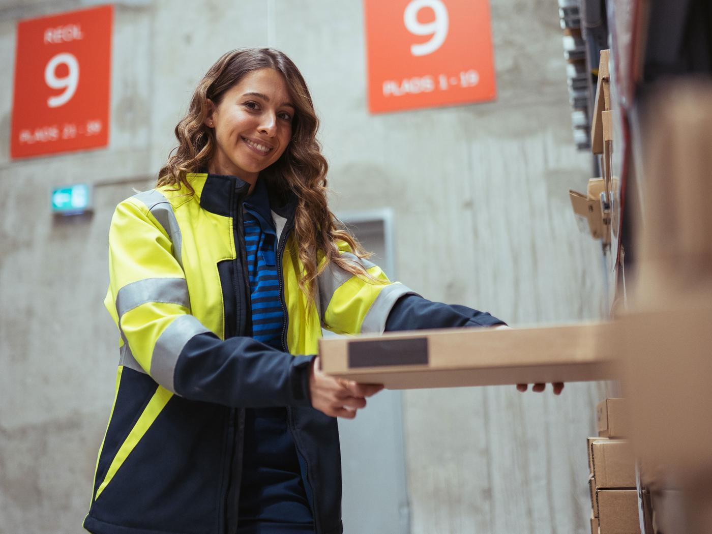 En person, der bærer en synlig jakke, smiler og holder en pakke på et lager, repræsenterer værdien af teamwork og effektivitet. Nummererede skilte med "9" er synlige i baggrunden.