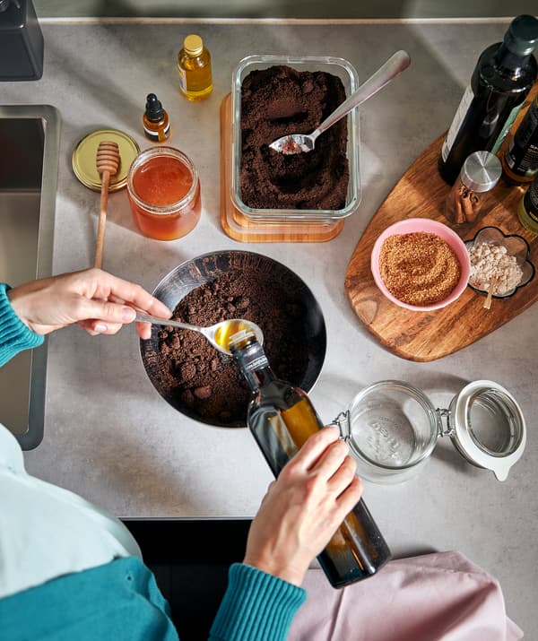 En person er i gang med at forberede en blanding ved et køkkenbord og hælder olie over en skål med kaffegrums. Ved siden af står et tomt KORKEN glas med låg.