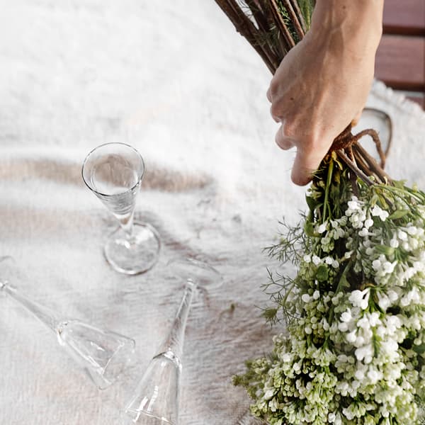 En person håller en bukett blommor ovanför en vit duk och bredvid ligger tre snapsglas.
