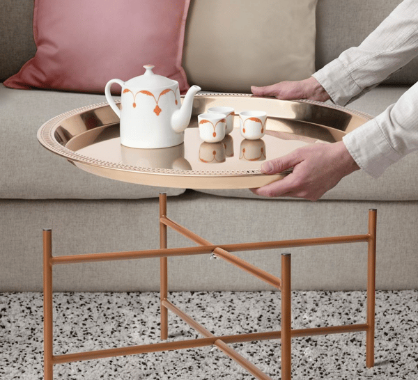 En person håller en rund bordsbricka med en tekanna och tre små koppar, ovanför ett bordsben i trä. I bakgrunden syns en soffa med rosa och beige kuddar.