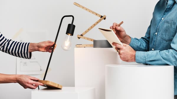 En person i en blå jeansskjorta skriver i en anteckningsbok och en annan person håller fram en TVÄRHAND bordslampa i svart/bambu.