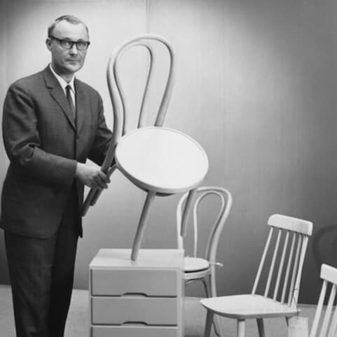 En person i svart kostym bland en scen med äldre möbelprototyper i ett utställningsrum, inklusive stolar och bord. 
