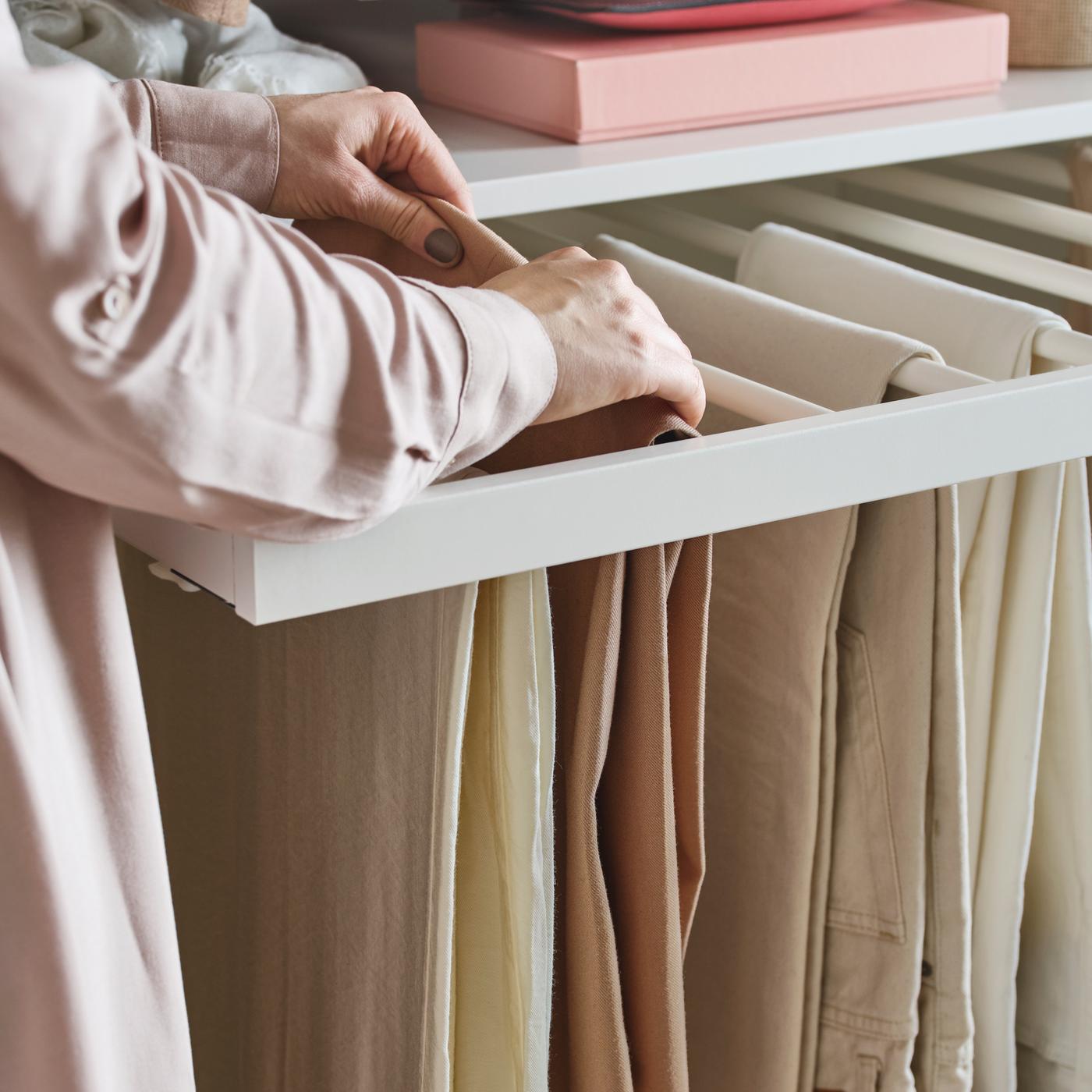 En person iført en pink bluse retter på et af flere par beige bukser, der hænger på en hvid KOMPLEMENT udtræksbuksebøjle.