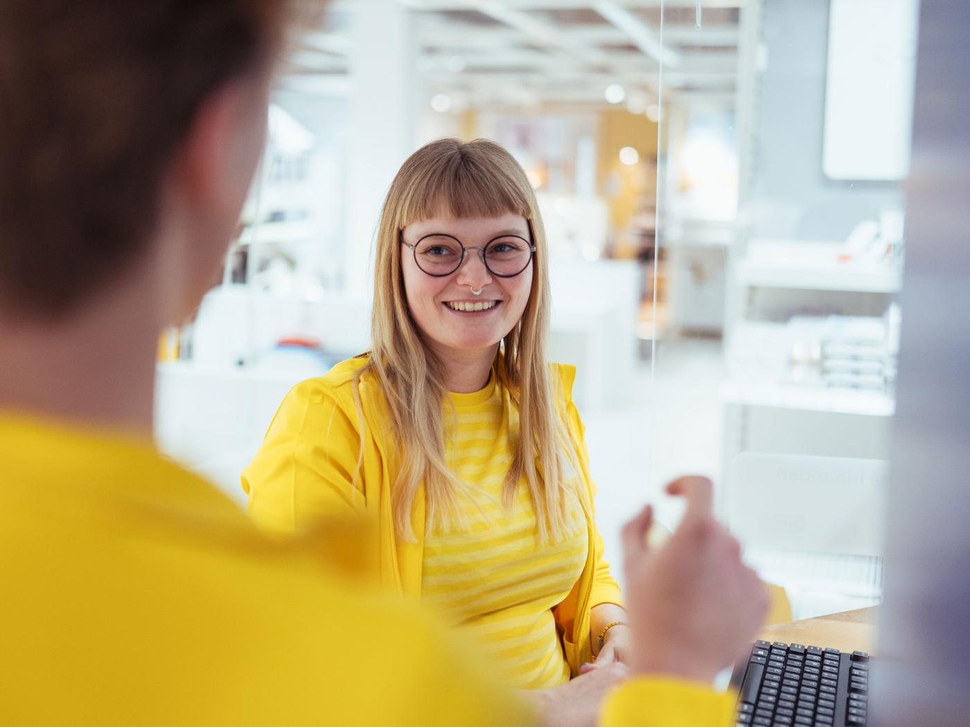 En person med langt blondt hår iført briller og et gult outfit smiler, mens han sidder ved et skrivebord på et lyst IKEA-kontor. En anden person, der også er klædt i gult, er delvist synlig i forgrunden, mens hun går i samtale på deres elevplads.