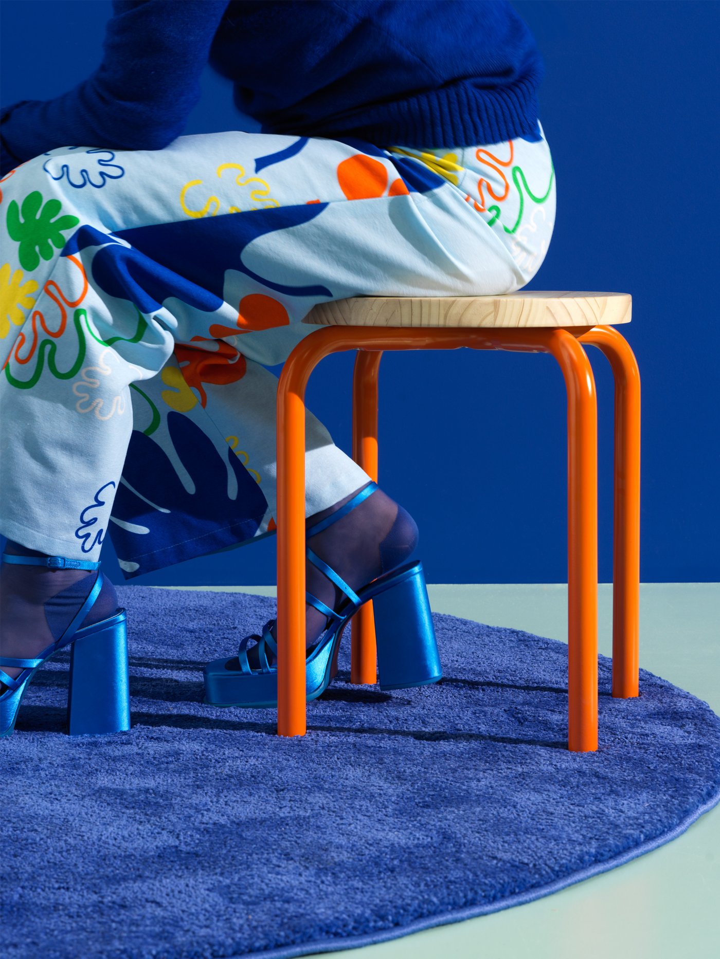 En person med mønstrede bukser sidder på en NYTILVERKAD taburet med orange ben ovenpå et blåt tæppe.