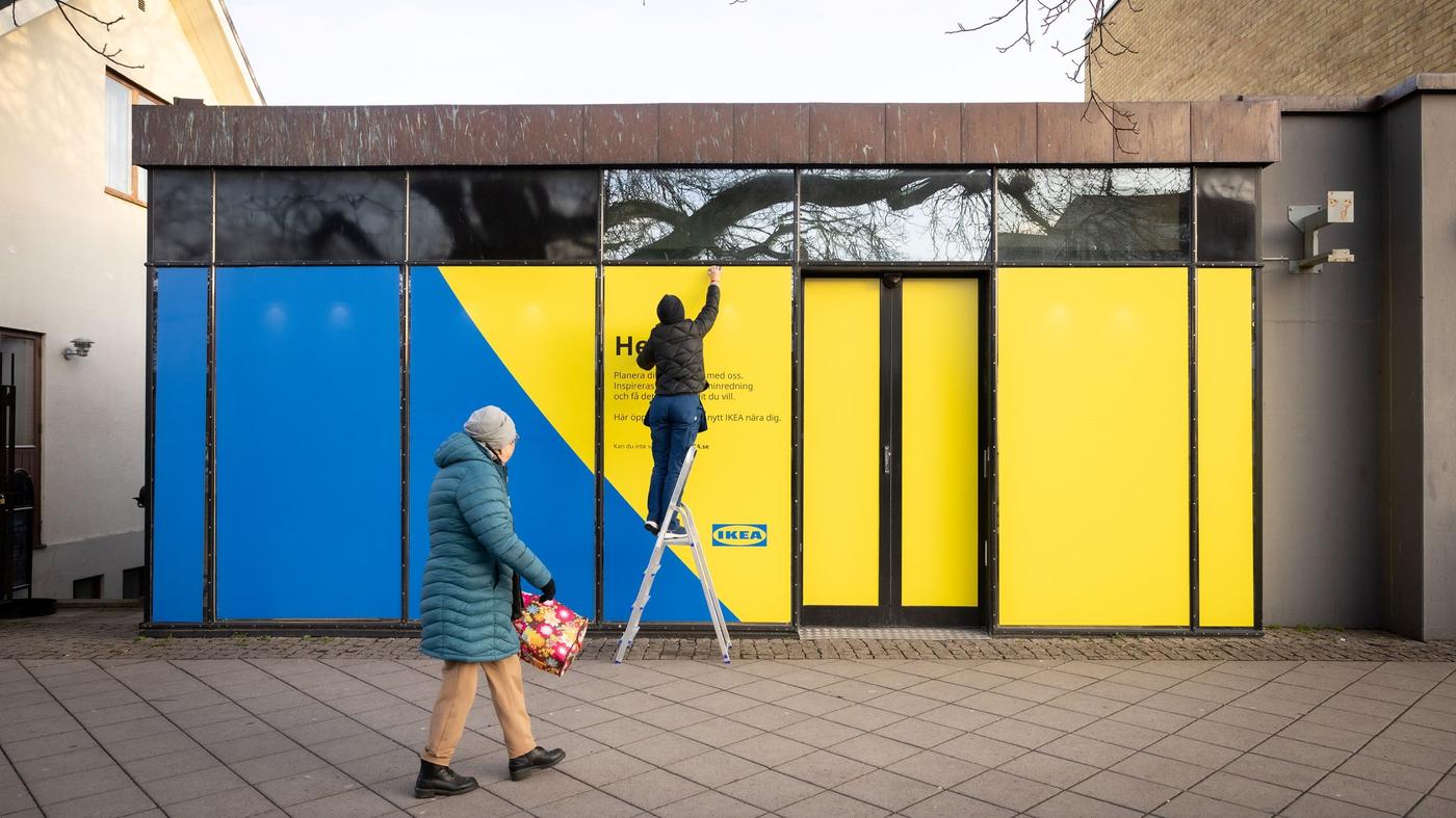 En person på stege som målar ett fönster på en byggnad i gula och blå färger. En annan person går förbi på trottoaren.