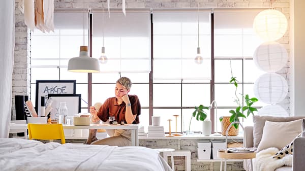 En person sidder ved et hvidt LAGKAPTEN/ADILS skrivebord under en BUNKEFLO loftlampe med et hvidt LACK sidebord bag sig.