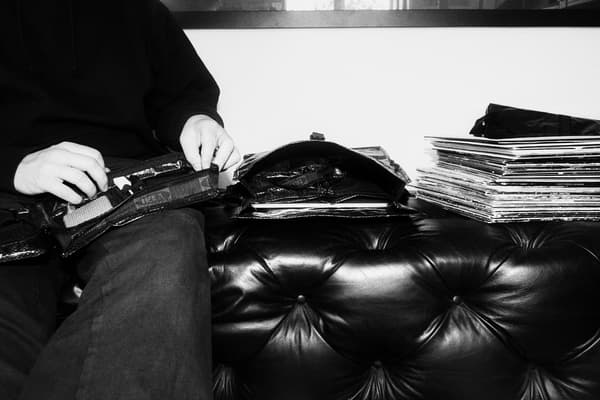 En person sitter på en skinnsoffa och fixar med en väska. Bredvid ligger ytterligare en väska och en hög med vinylskivor.